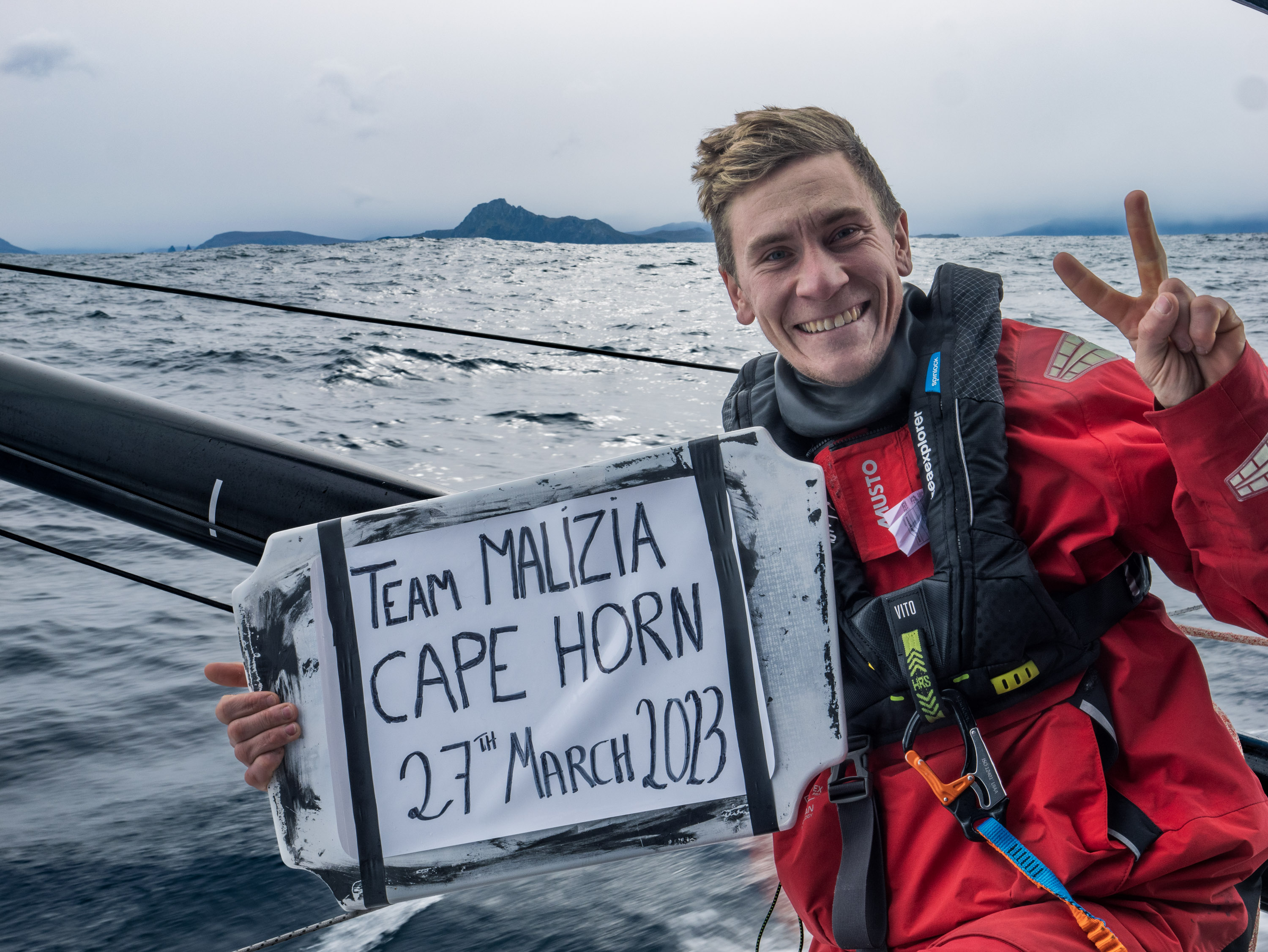 Sailor in red offshore gear at sea holding a handwritten Cape Horn sign dated March 2023