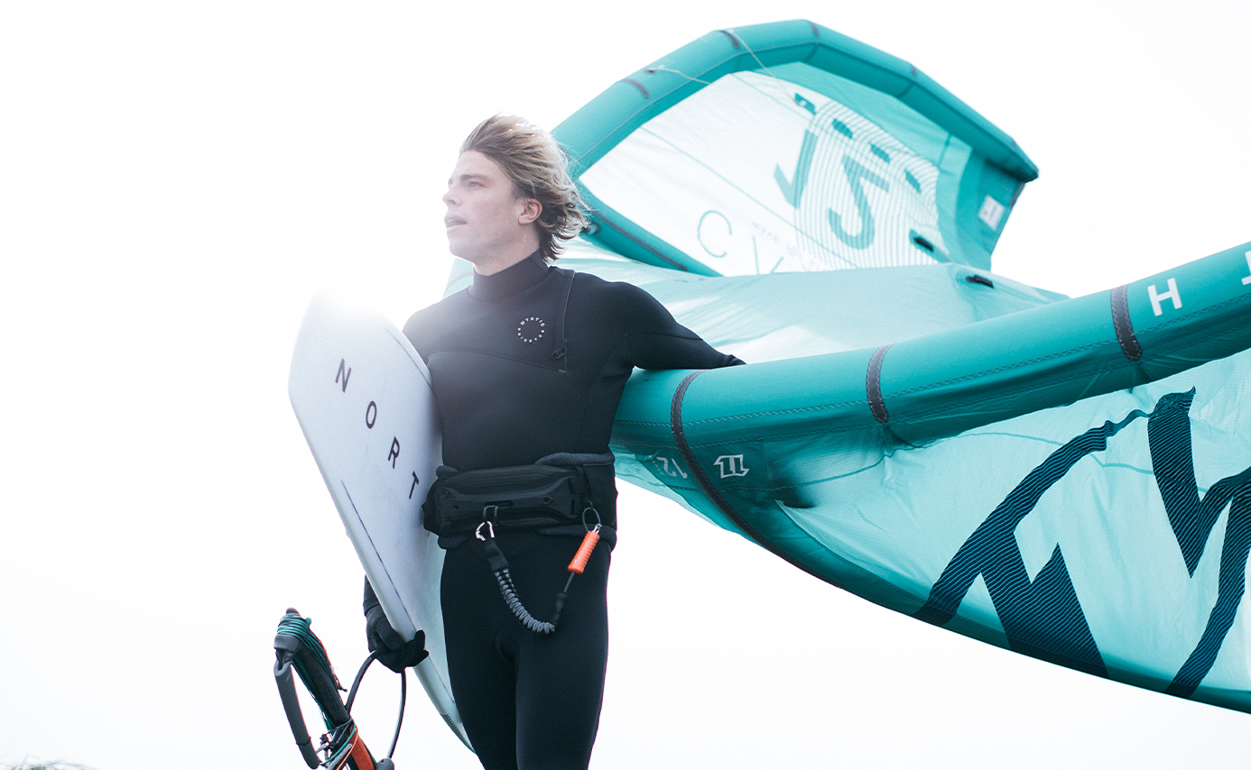 Kitesurfer im schwarzen Neoprenanzug mit weißem Board und türkisem Tubekite vor weißem Hintergrund