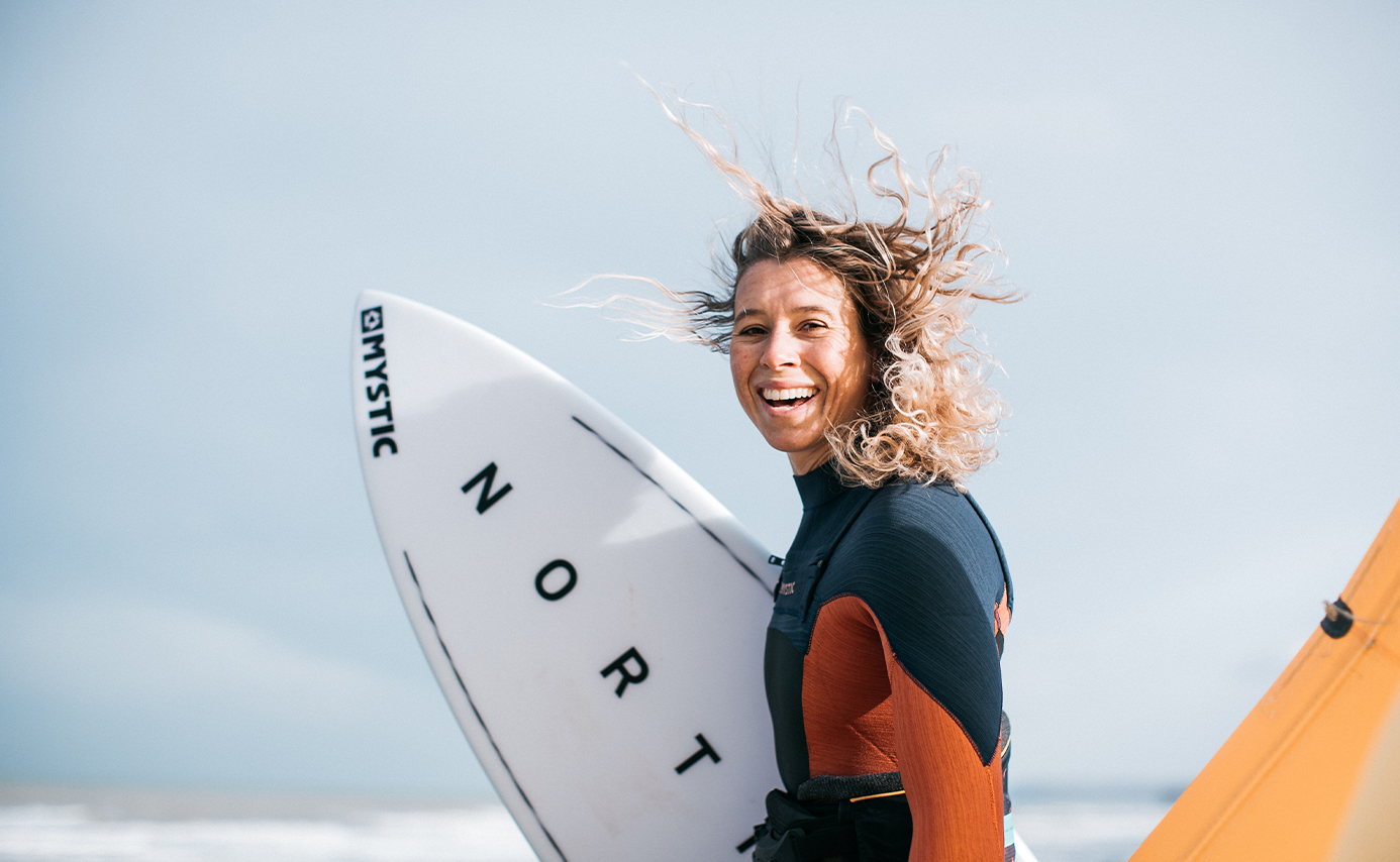 Lächelnde Frau mit windzerzausten Haaren trägt ein weißes North Board, in Neoprenanzug und orangem Trapez